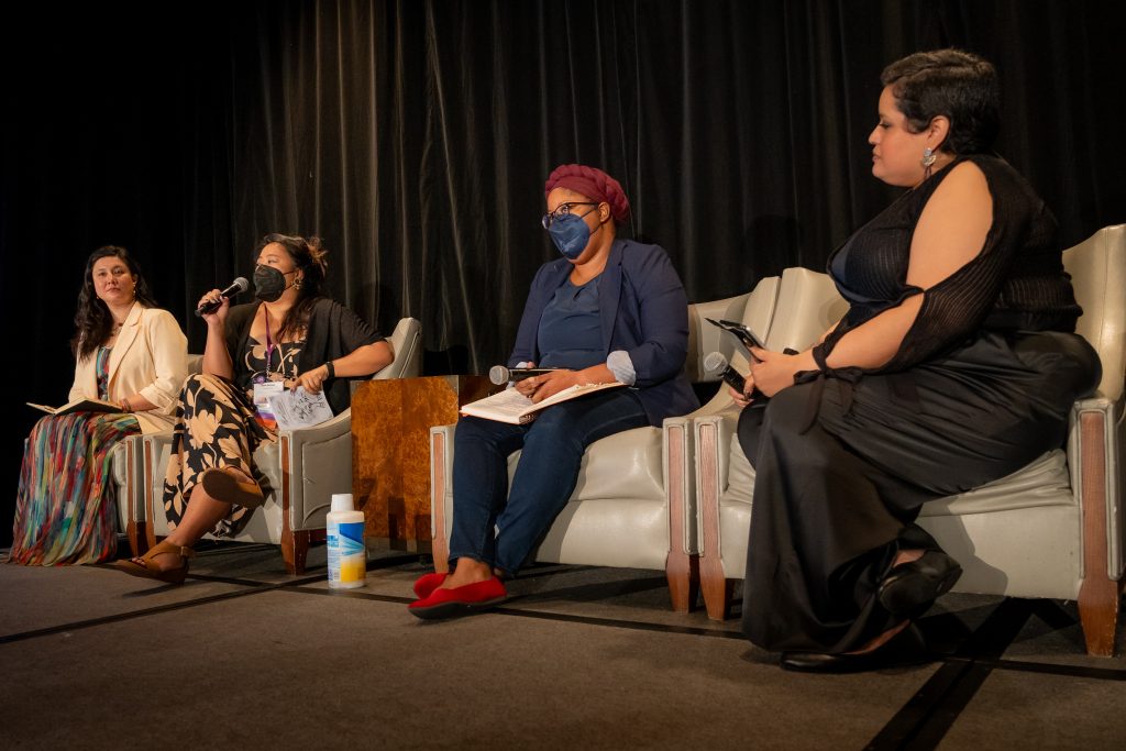 Four brown and Black women sit on a stage in white chairs. Two wear masks, one holds a mic. They are turned to each other. Each woman is an expert in reproductive and racial justice organizing: "AnaYelsi Velasco Sanchez, Jennifer Driver Elaina Ramsey, and Isabelle Leighton. AnaYelsi Velasco Sanchez, Jennifer Driver Elaina Ramsey, and Isabelle Leighton.