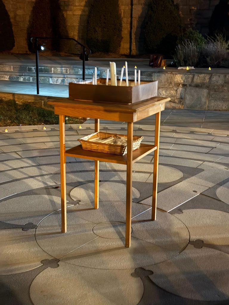 Four legged wooden table in the middle of a stone labryinth. On the top is an open box of sand with several narrow while candles standing in it. Some are lit, some are not. A box of unlit candles sits on a shelf below.